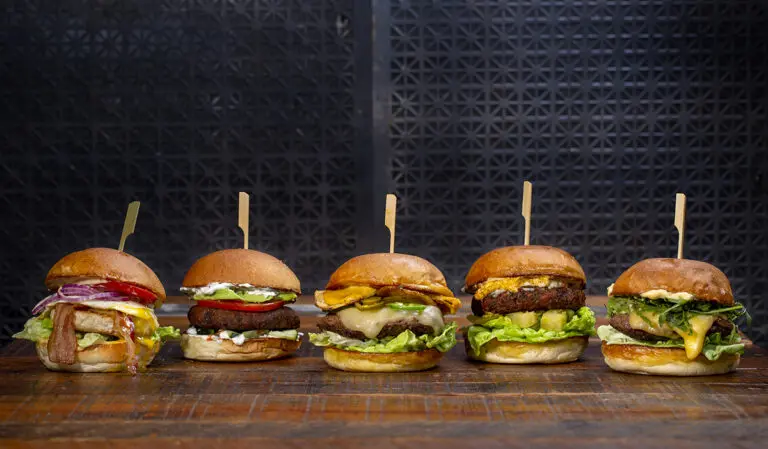 A row of five cheeseburgers on a wooden table against a black metal background.