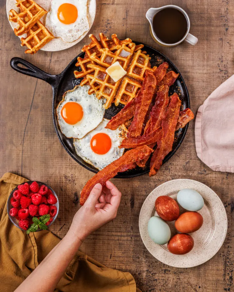 Photo of a hand reaching in to grab a piece of bacon from a family style breakfast serving.