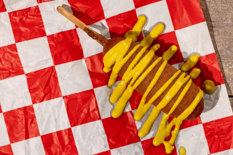 Photography of a picnic scene on a wooden table with a corndog covered in mustard on a red checkered napkin.