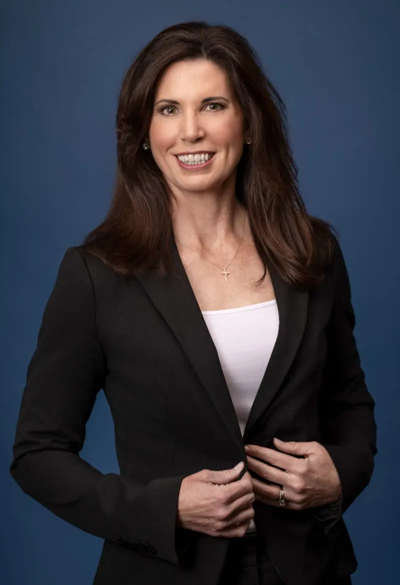 Corporate headshot of a woman in a black blazer against a blue studio backdrop, photographed by Beth Hall Photography in Fayetteville, Arkansas.
