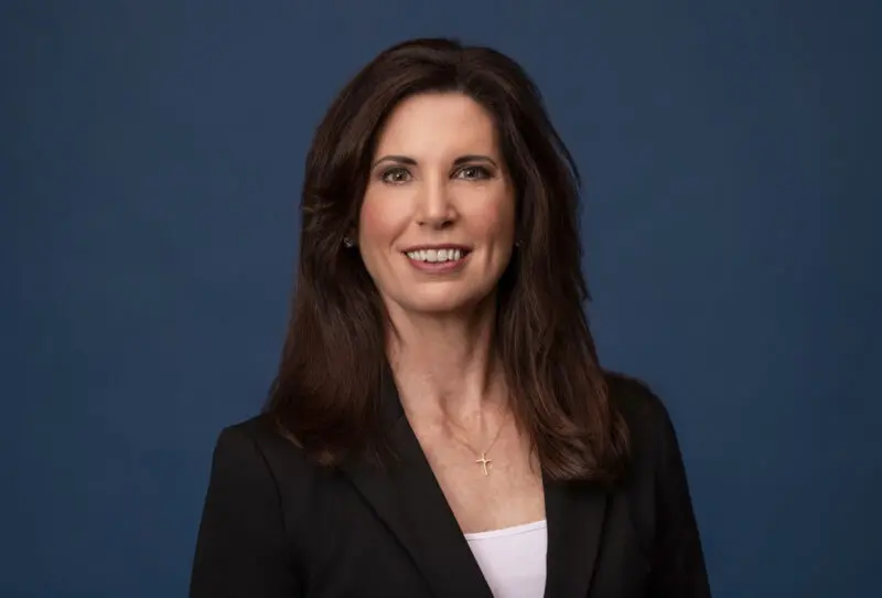 Corporate headshot of a woman in a black blazer against a blue studio backdrop, photographed by Beth Hall Photography in Fayetteville, Arkansas.