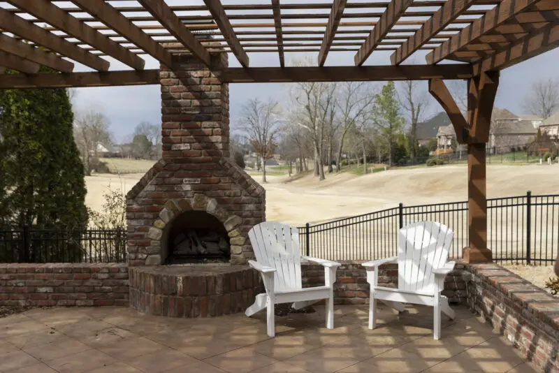 Two white adirondak chairs under a pergola on a porch.