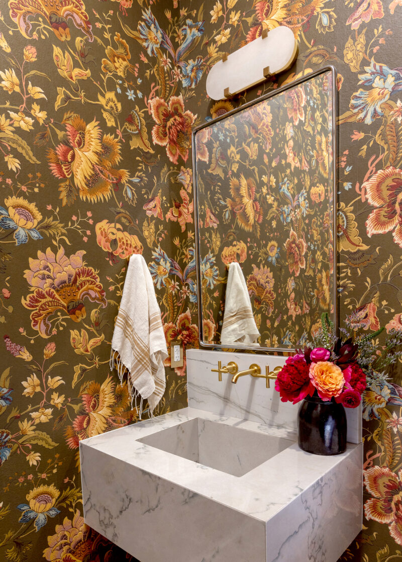 A powder bathroom with decorate floral wallpaprt in browns, golden tones and reds decorated with a vase of peonies.