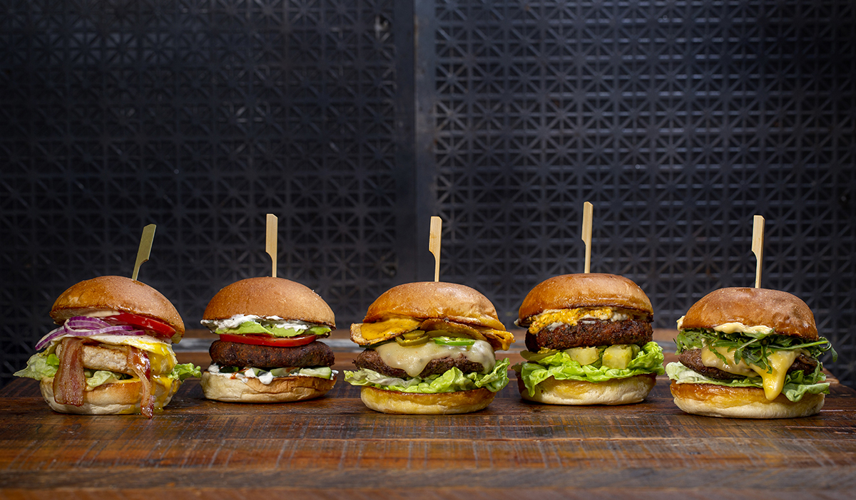 A row of five cheeseburgers on a wooden table against a black metal background.
