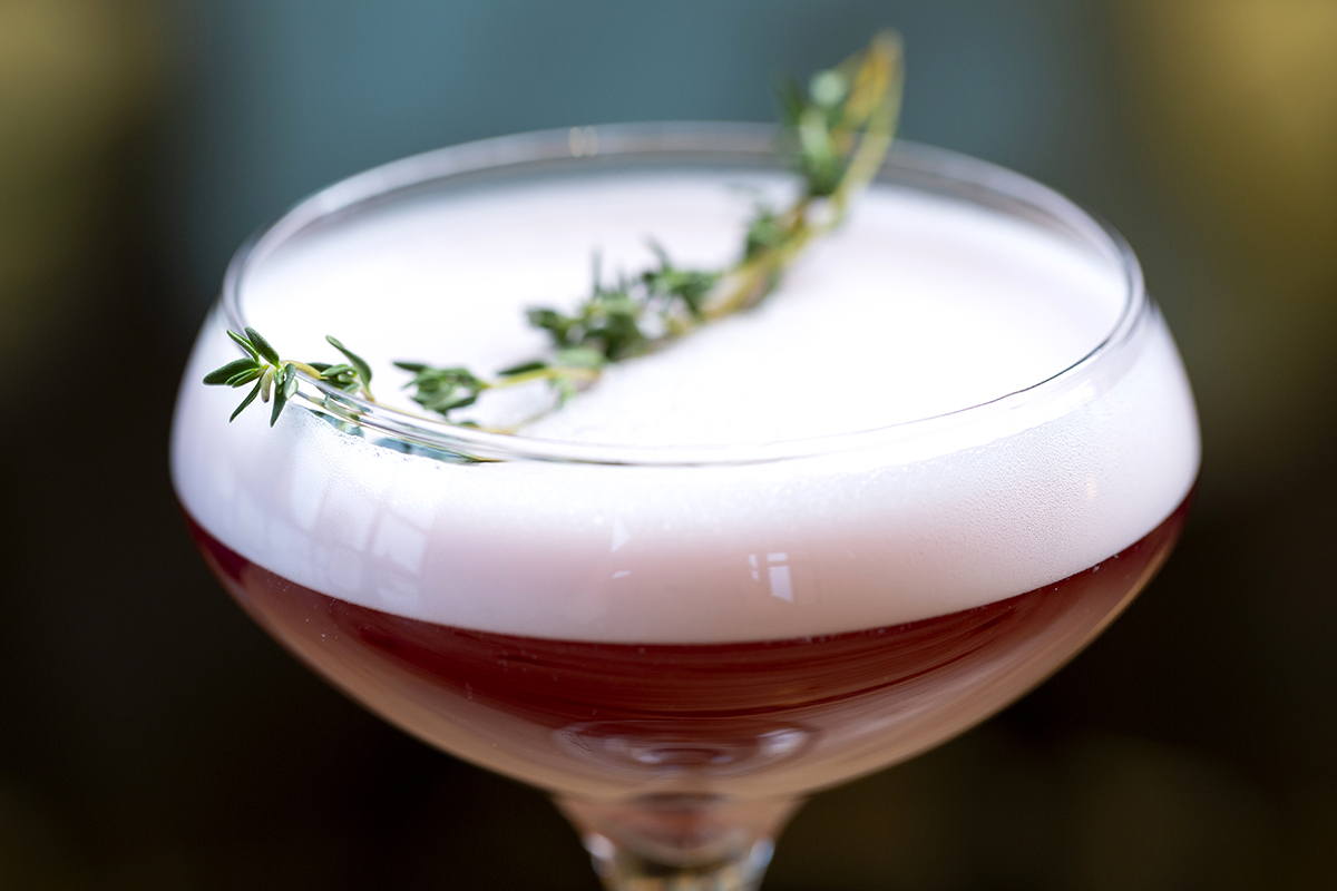 Close up photography of a maroon drink with a spring of thyme as garnish.