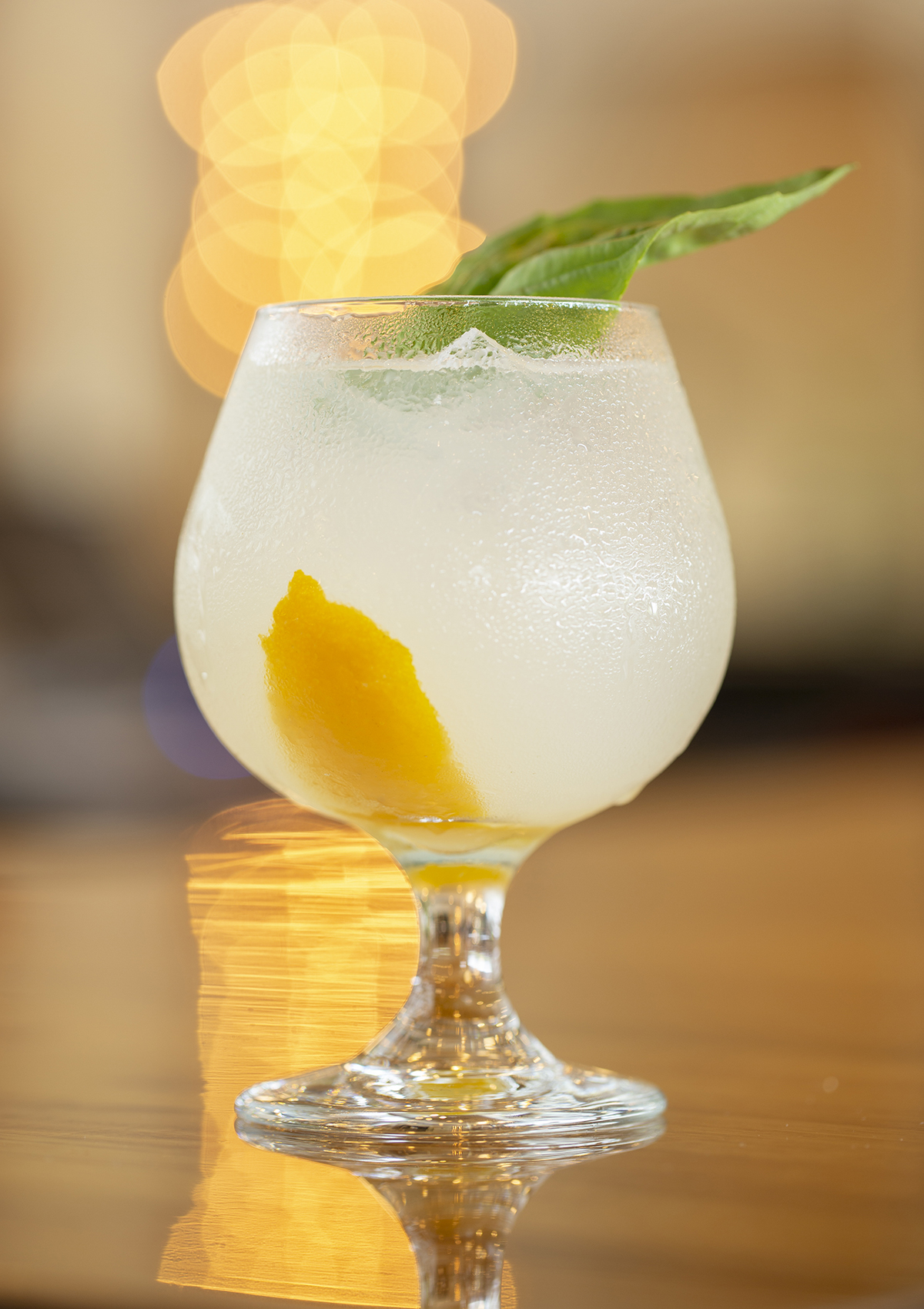 A pale yellow drink on a bar with a large basil leaf for garnish.