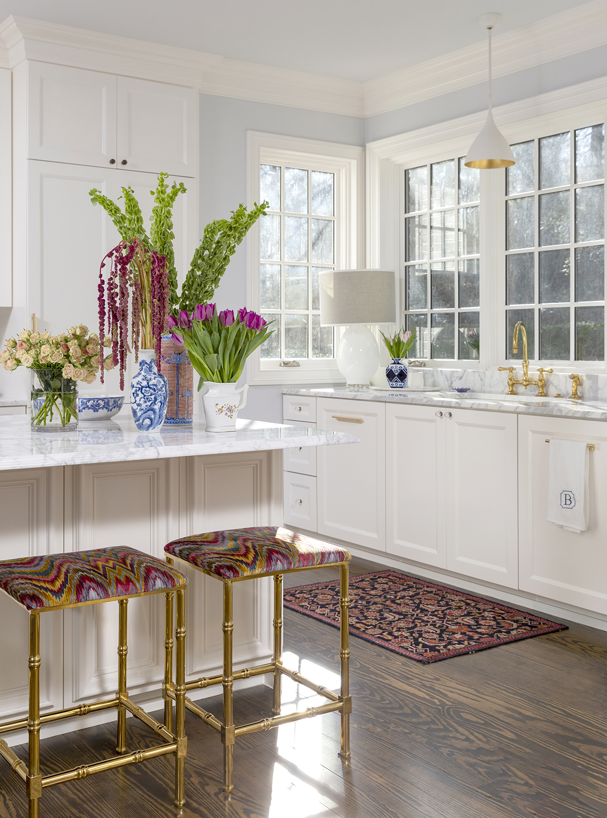 Interior photography of a white kitchen with gold stools, colorful fabric and flowers and a rug in a home in Fayetteville, Arkansas.