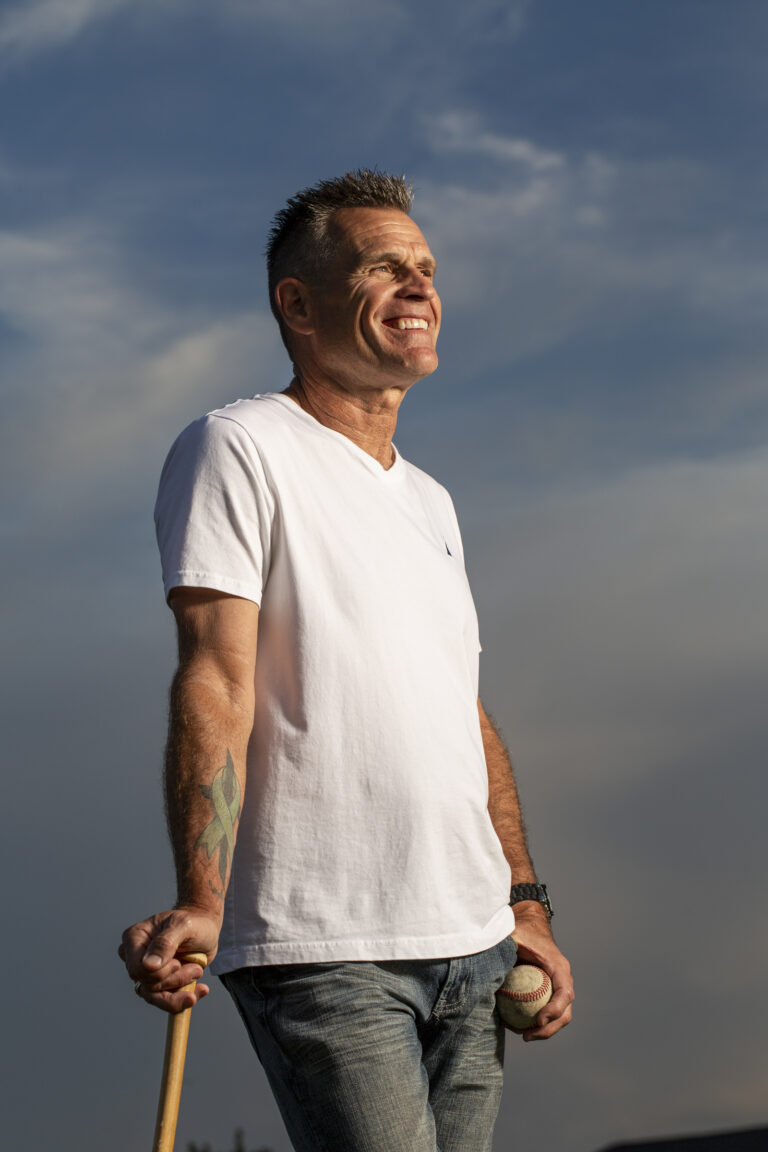 A portrait of a man posing against a blue sky while holding a baseball