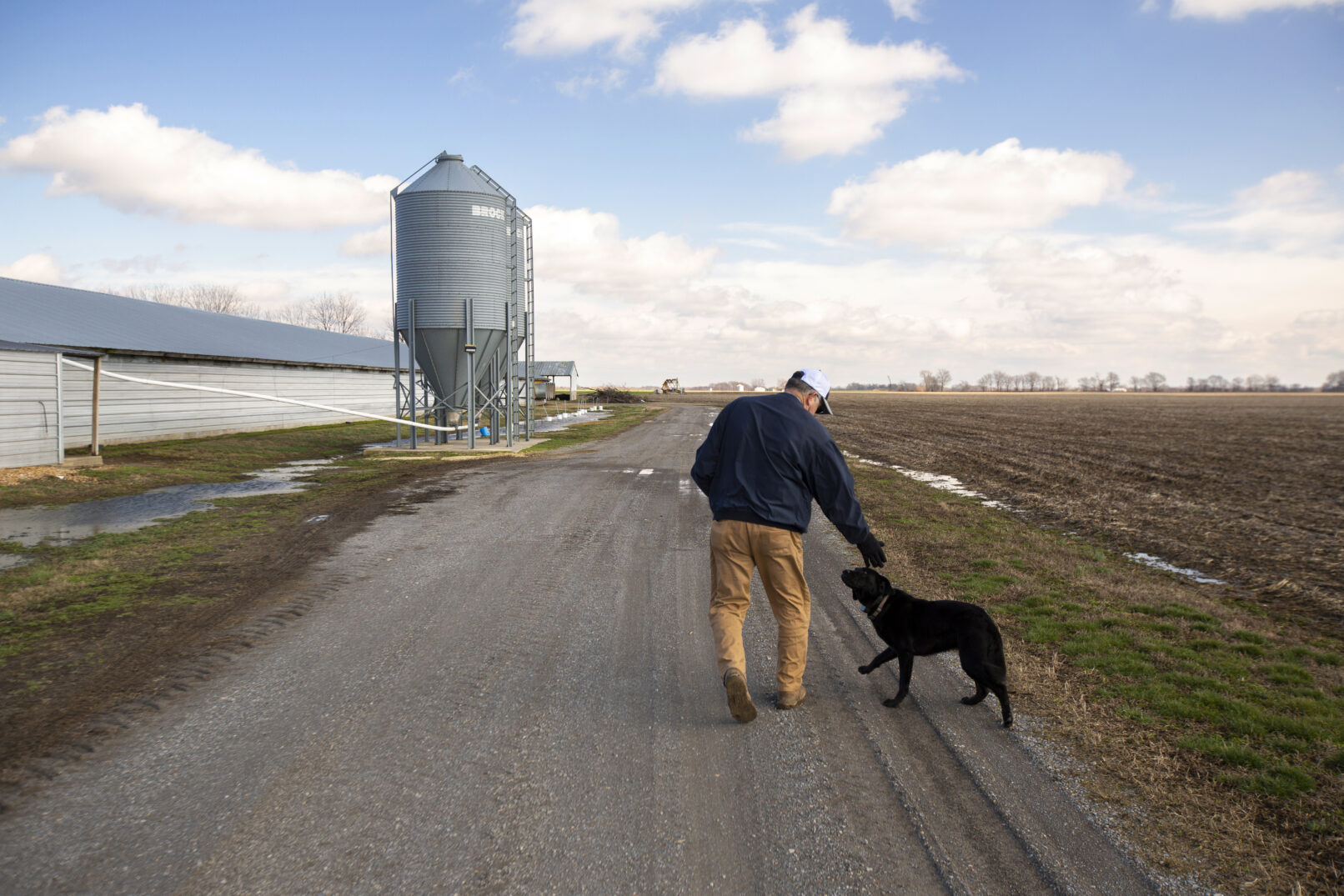 Lifestyle portrait of a farmer walking down a gravel road petting his dog.