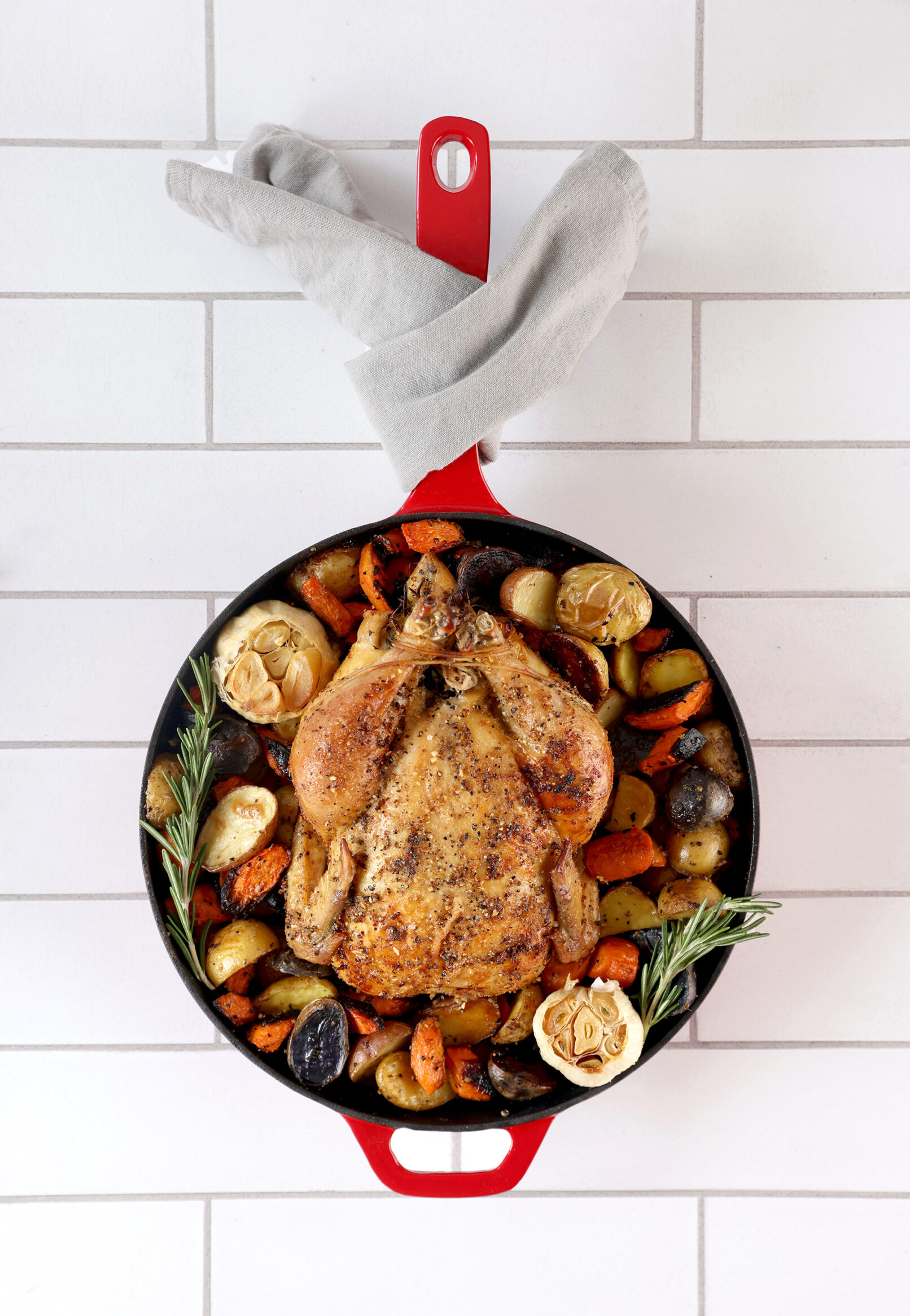 Photo of a whole, roasted chicken in a red Le Creuset skillet on a white tile background.