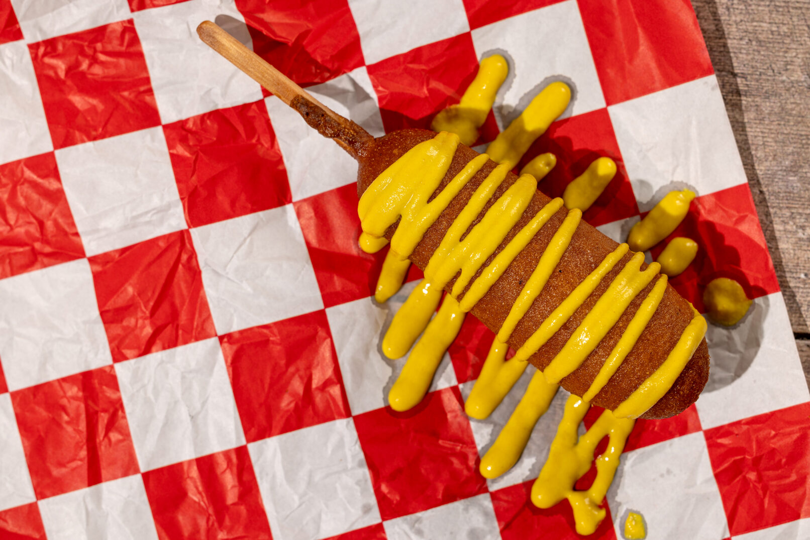 Photography of a picnic scene on a wooden table with a corndog covered in mustard on a red checkered napkin.