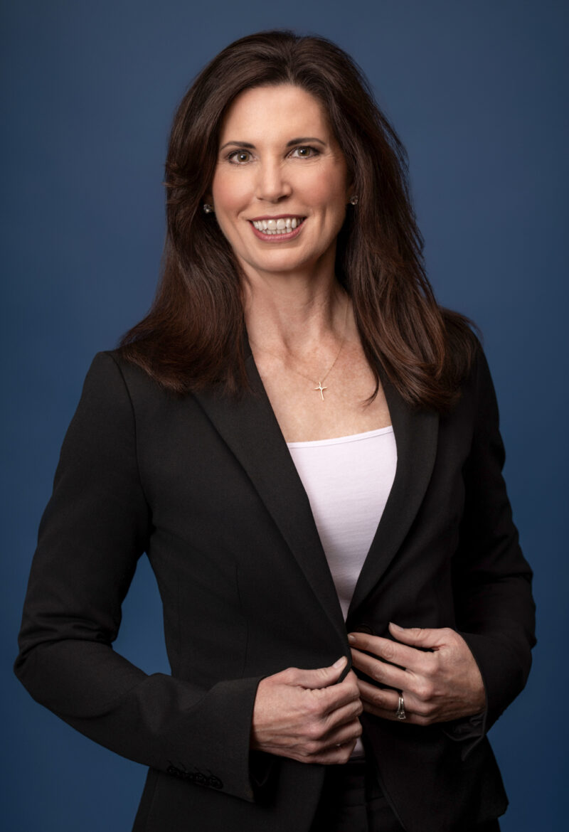 Corporate headshot of a woman in a black blazer against a blue studio backdrop, photographed by Beth Hall Photography in Fayetteville, Arkansas.