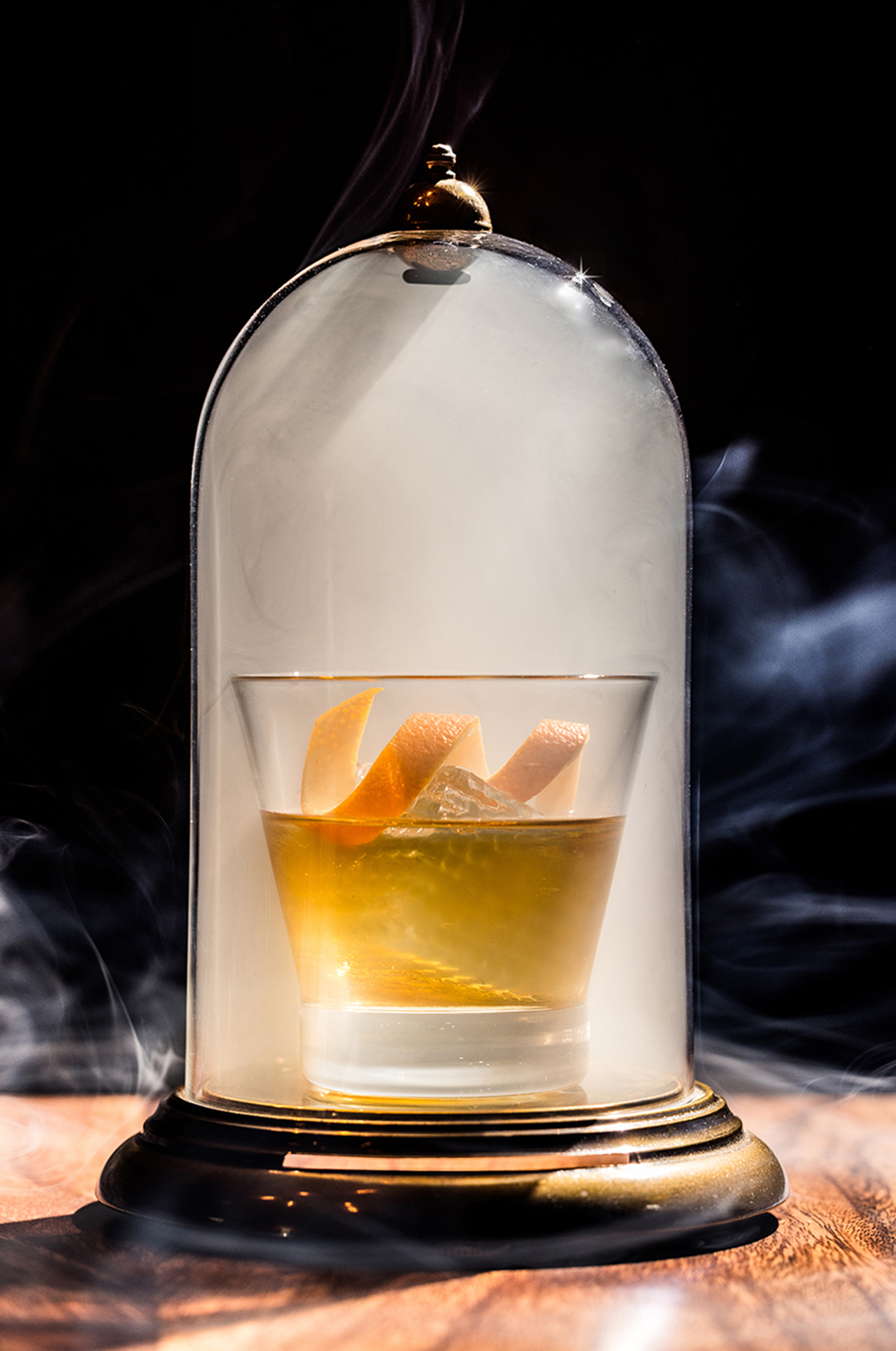 Photo of a smoked old fashioned with a twisted orange garnish under a glass dome on a wood table.