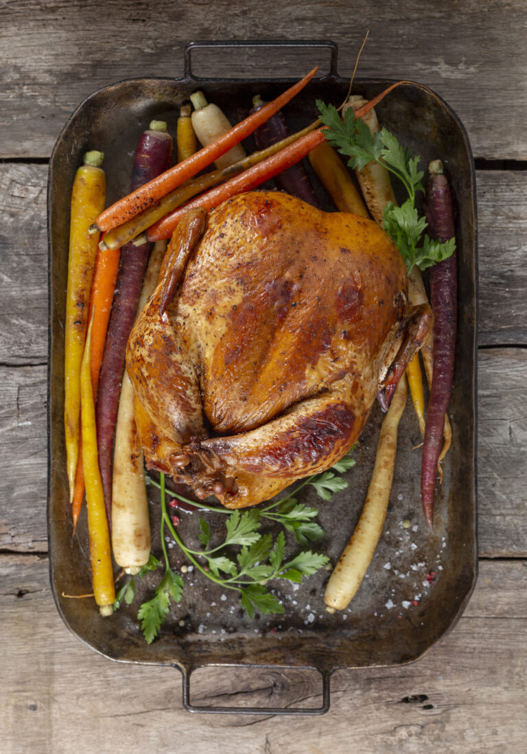 Photo of a whole, cooked chicken on a metal pan with roasted carrots.