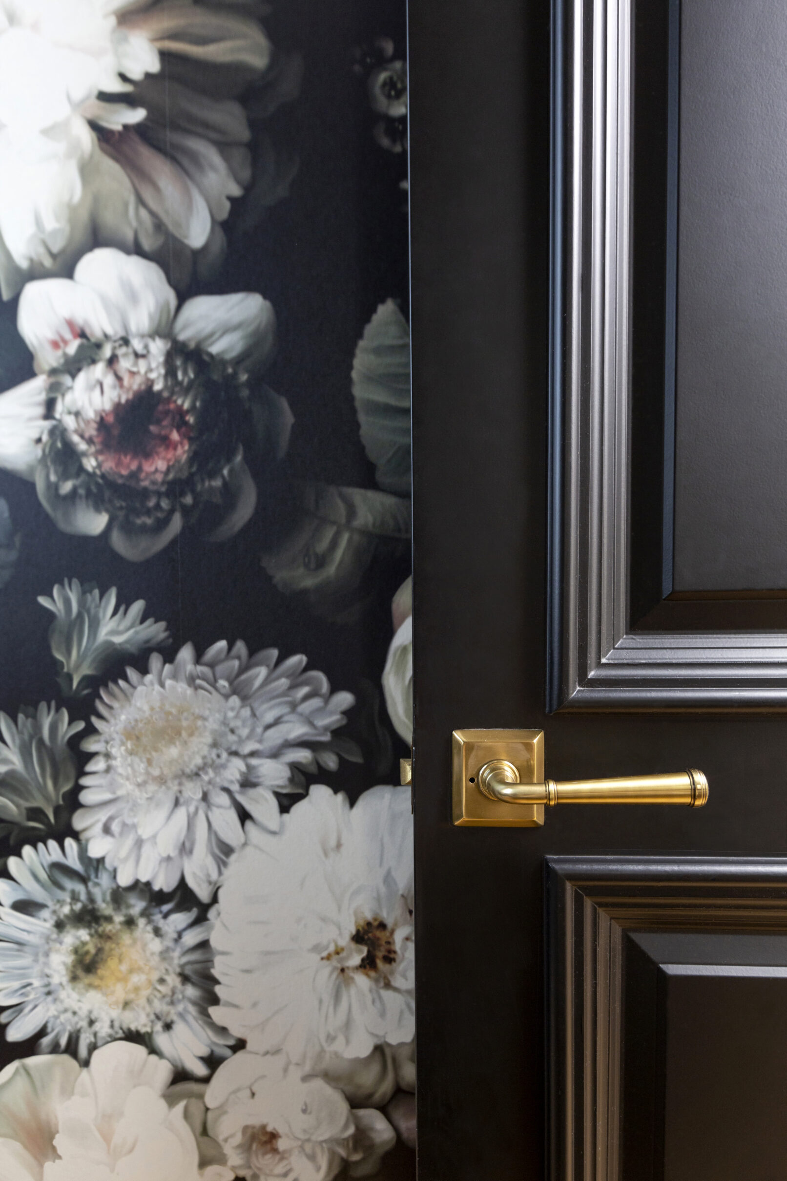 Detail photo of a black door with a gold handle in front of floral wallpaper.