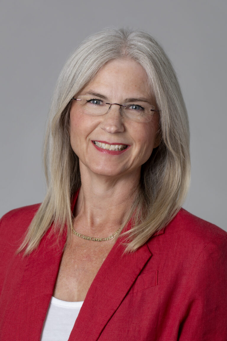 Studio headshot of a woman in a read jacket in front of a gray background.