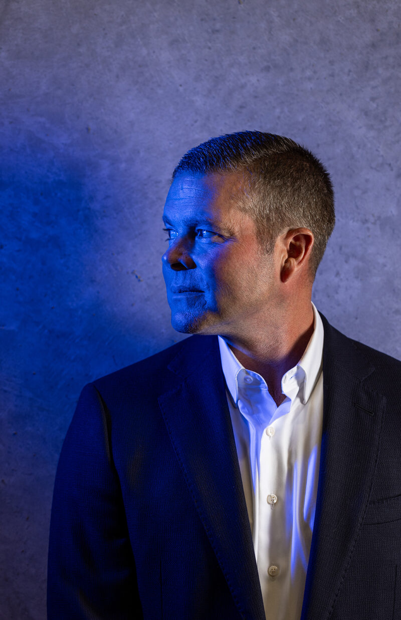 An executive stands near a concrete wall and has a blue light on his face.