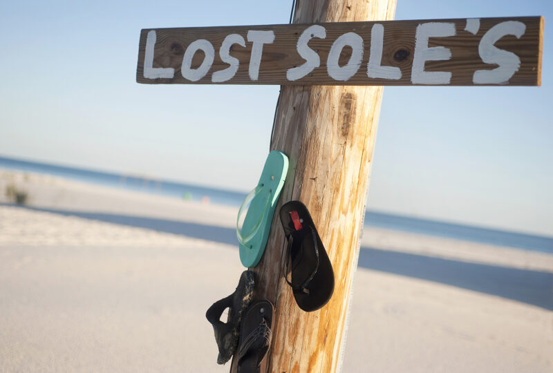 A wooden sign on a beach with the wors lost soles and single flip flops hanging on a post.