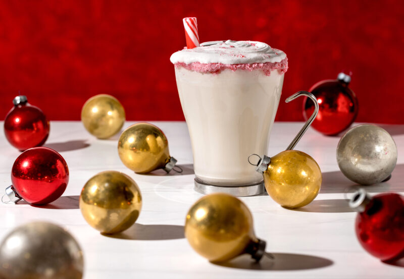 Spiked Eggnog with peppermint on the rim of a clear glass. The glass is surrounded by red, gold and silver ornaments in front of a bright red background.